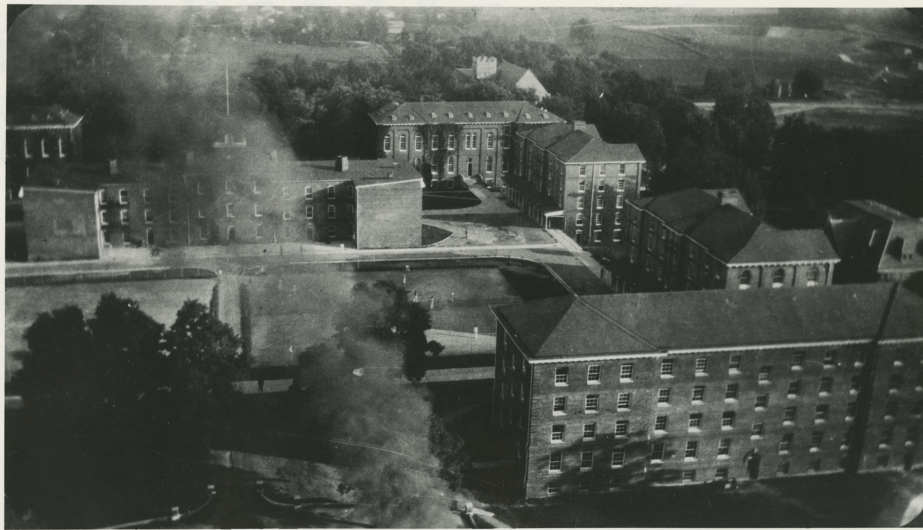 "Looking south from the tall smokestack of the new powerplant. Barracks No. 1 at left center. Clockwise around Barracks No. 1, starting at the top left: First Academic Building, Second Academic Building, Barracks No. 3 [part of the former Brodie Hall], Barracks No. 5, Barracks No. 6." You can also see part of the old library at the top center. 