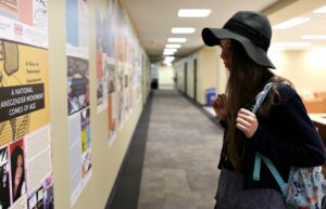 Photograph of a female-presenting person wearing a blue open knit sweater and black felt cloche hat holding a light blue backpack. This person is viewing the exhibit posters displayed on a wall.