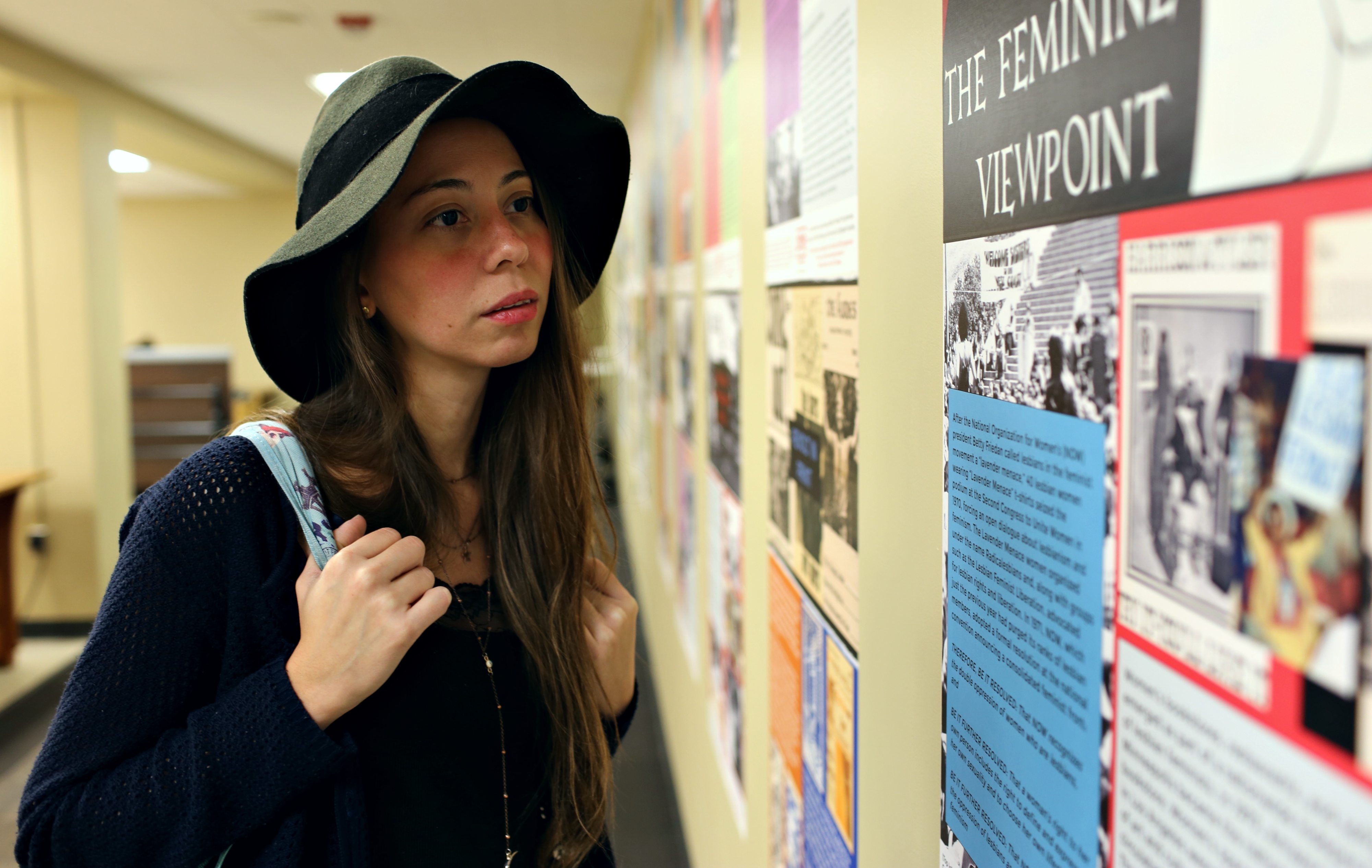Photograph of a female-presenting person wearing a blue open knit sweater and black felt cloche hat holding a light blue backpack. This person is viewing the exhibit posters displayed on a wall.