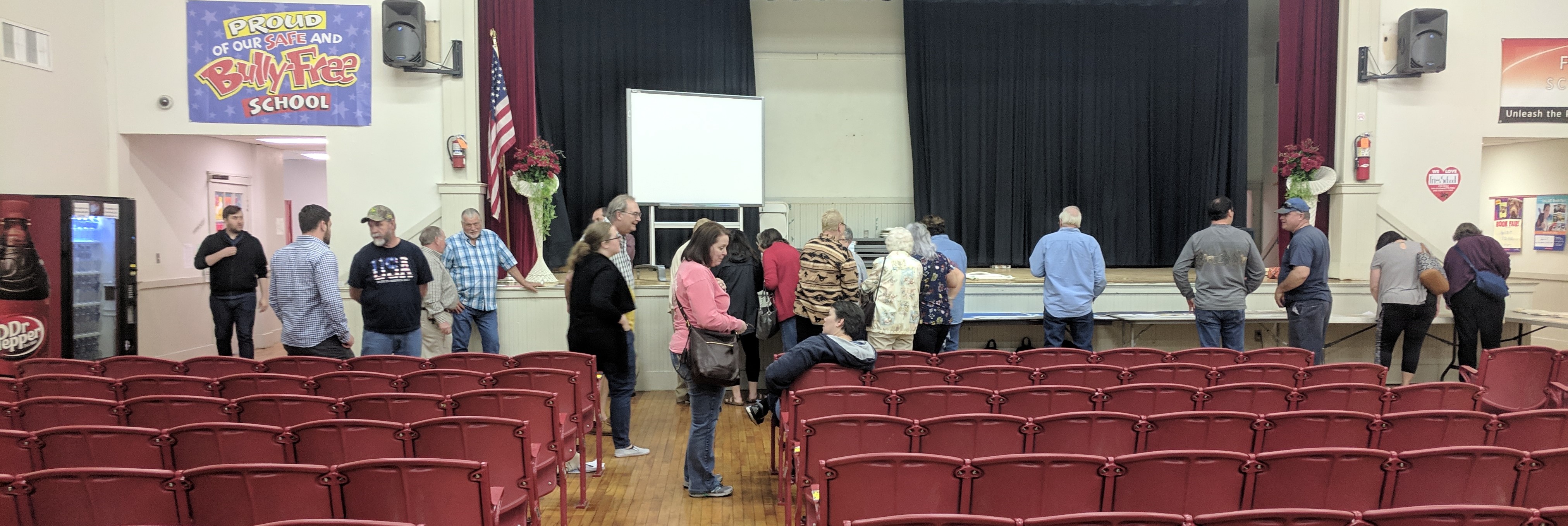 A line of people in front of the stage in a middle school auditorium talk to each other and look at copies of historical documents. Red auditorium seating fills the foreground.