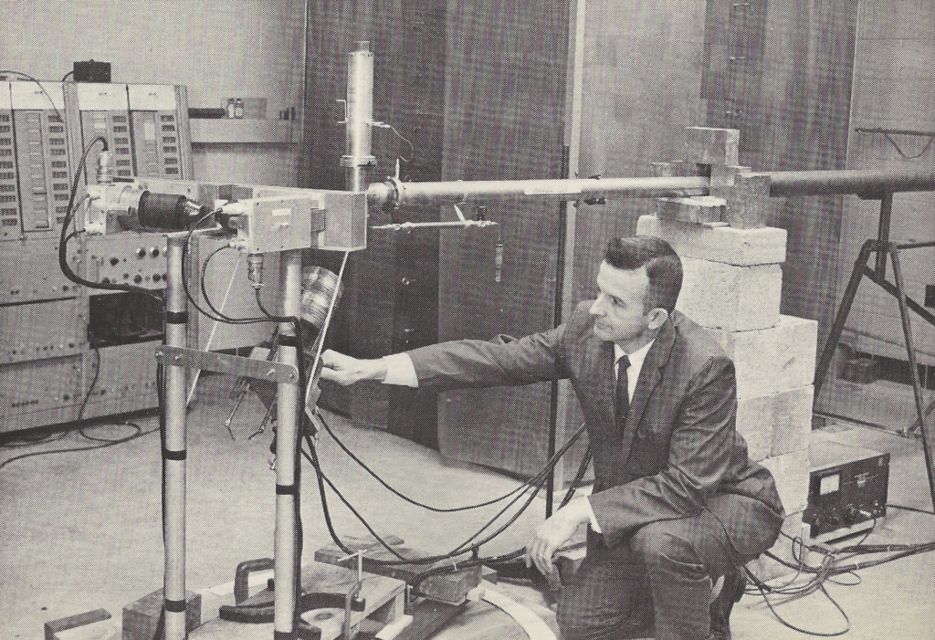 Black and white photo of a white man in a dark suit kneeling next to an assembly of metal tubes, wires, and concrete blocks.